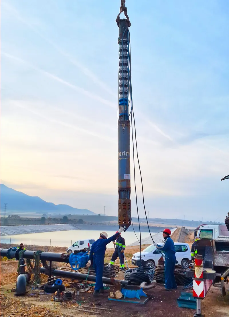Equipo VITTA suspendido de la grúa sobre la boca del pozo durante la maniobra de instalación, junto a una balsa de riego